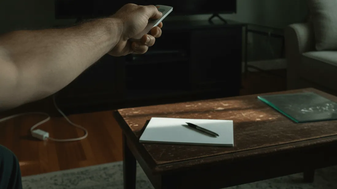 A hand holds a remote control pointed at a dark television screen in a dimly lit room with a coffee table nearby.