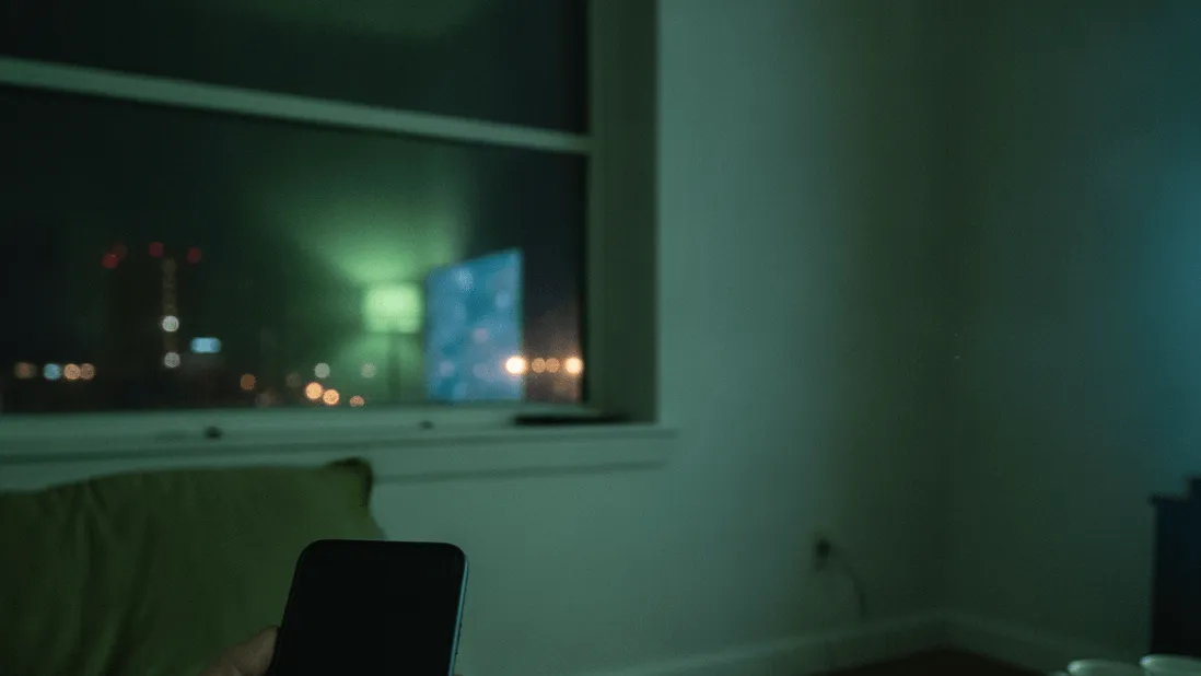 A hand holding a smartphone with a dark screen, a remote, and a window reflecting a TV glow at night.