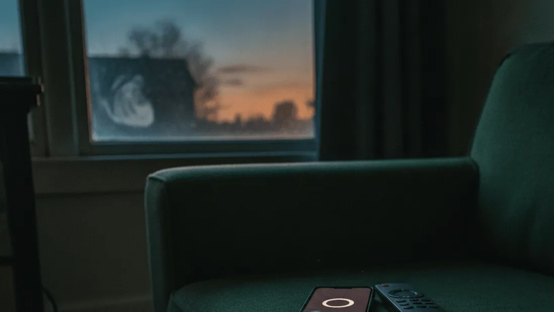 A smartphone displaying a blank loading circle rests on a couch armrest beside a remote control, at dusk.