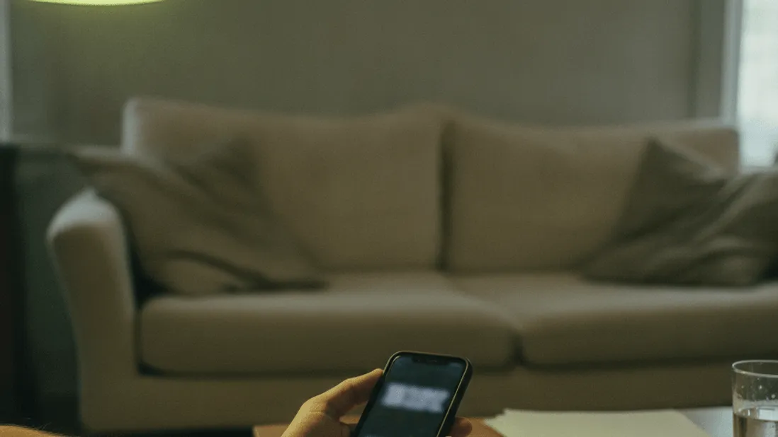 A hand holds a phone timer beside a notepad on a coffee table under warm lamp light.