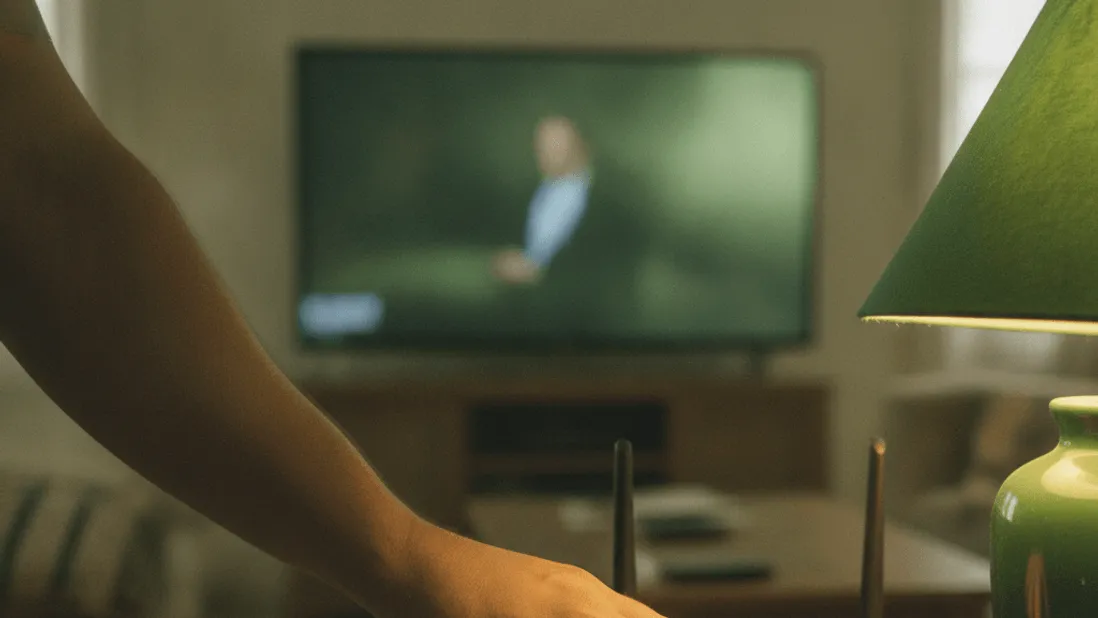 A hand adjusts a router on a wooden shelf next to a green lamp, with a blurred television in the background.
