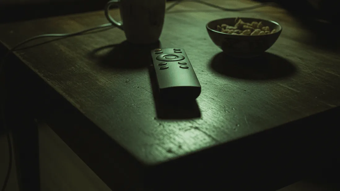 A dimly lit coffee table with a remote, a mug of tea, and a bowl of snacks, with a person on a couch.