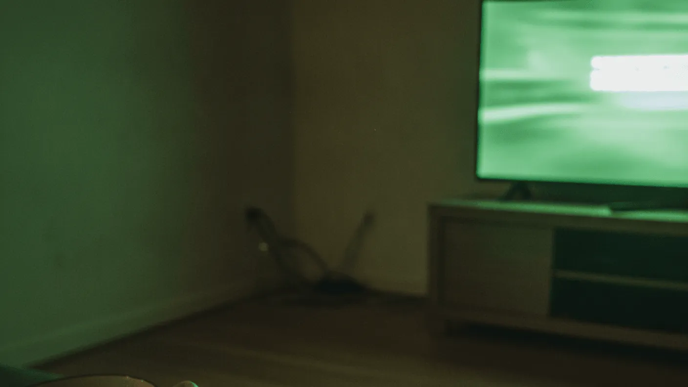 A remote and a hand rest on a couch armrest, with a bowl of snacks, illuminated by a green-tinted screen.
