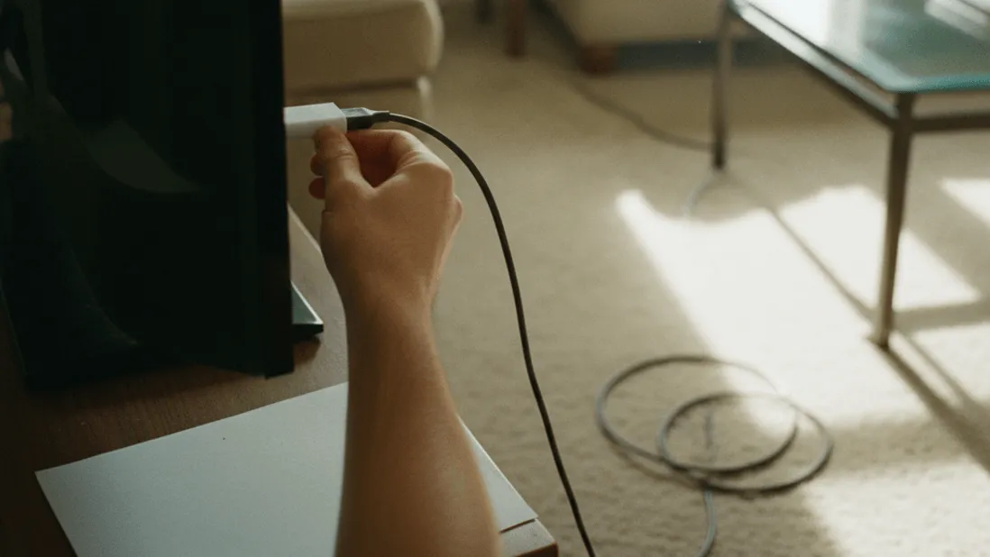 A hand adjusts an HDMI cable connected to a streaming stick behind a television, with sunlight on a dusty carpet.