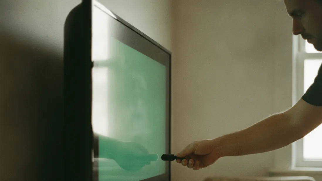 A person's hand adjusts a streaming stick in the back of a television with cables and a mug on a shelf below.