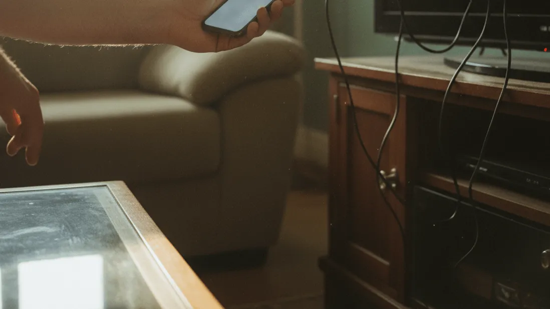 A man's hand holds a phone near a TV with a streaming stick, in a sunlit living room.