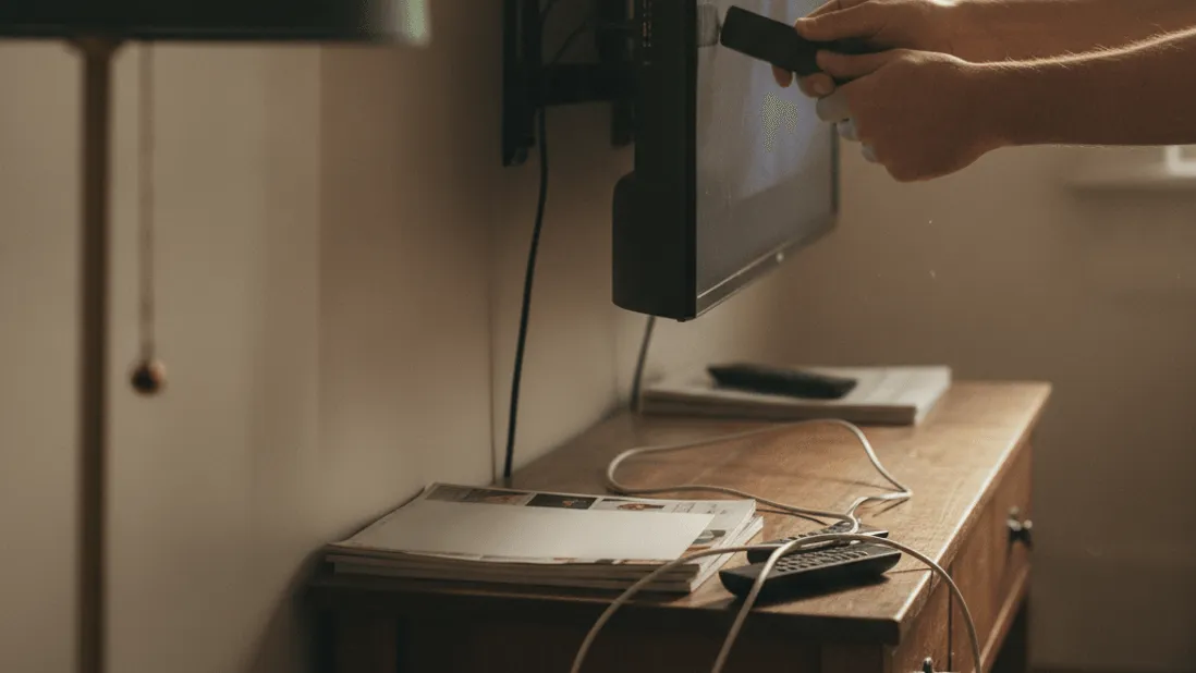A pair of hands plugs a streaming stick into the back of a wall-mounted television in a warm, lived-in room.