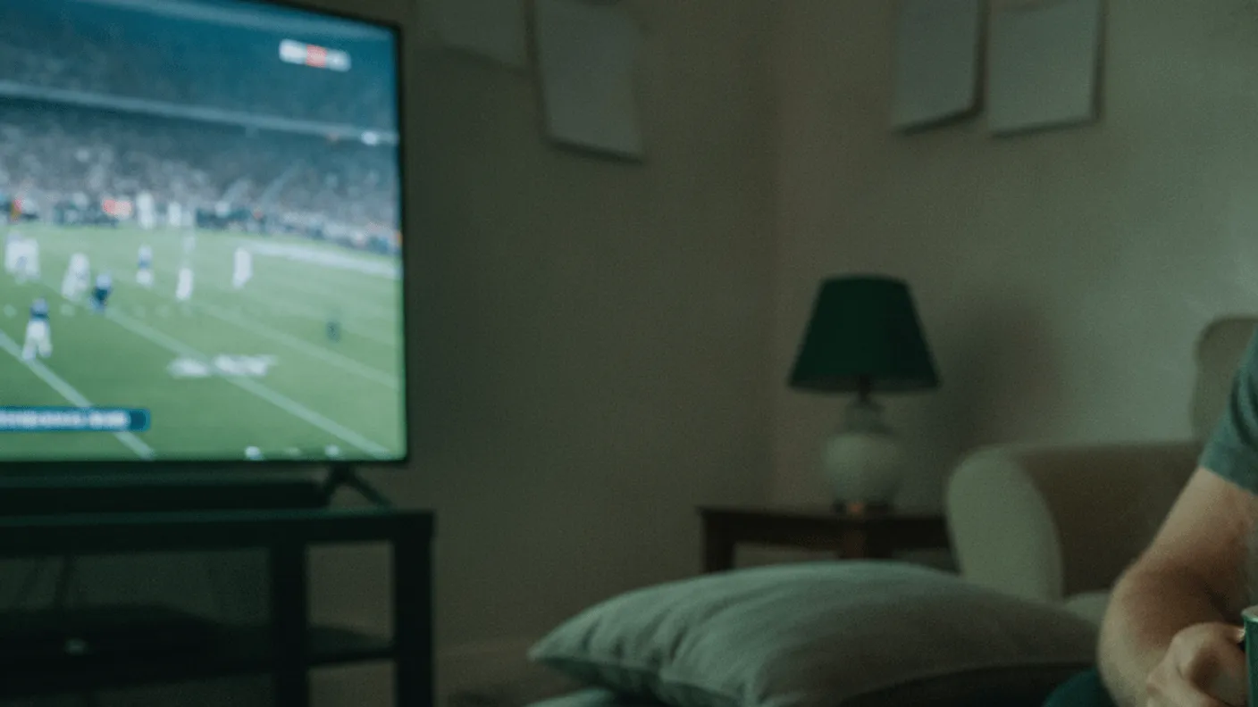 A remote control rests on a wooden coffee table in a dim room with a paused football game on TV.