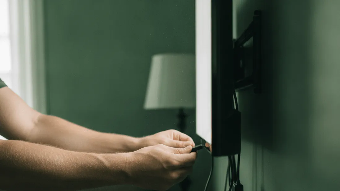 A person's forearms adjust a streaming stick plugged into a wall-mounted TV with tangled cables.