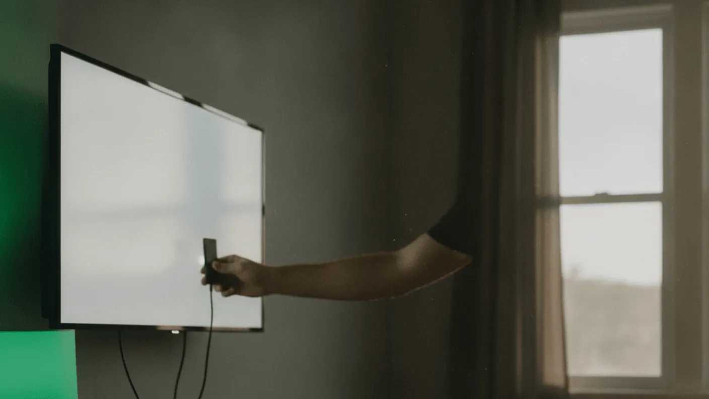 A man's arm inserts a device into a wall-mounted TV, with cables, a wooden shelf, and a window in the background.